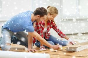 A man in blue shirt and woman in red and white plaid shirt measuring wood flooring for winter home improvement project.)