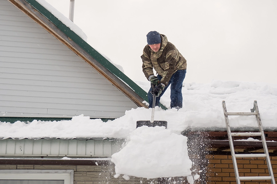 Man on his Springfield, IL roof with a ladder propped up against the house using a shovel to remove snow off of his roof.